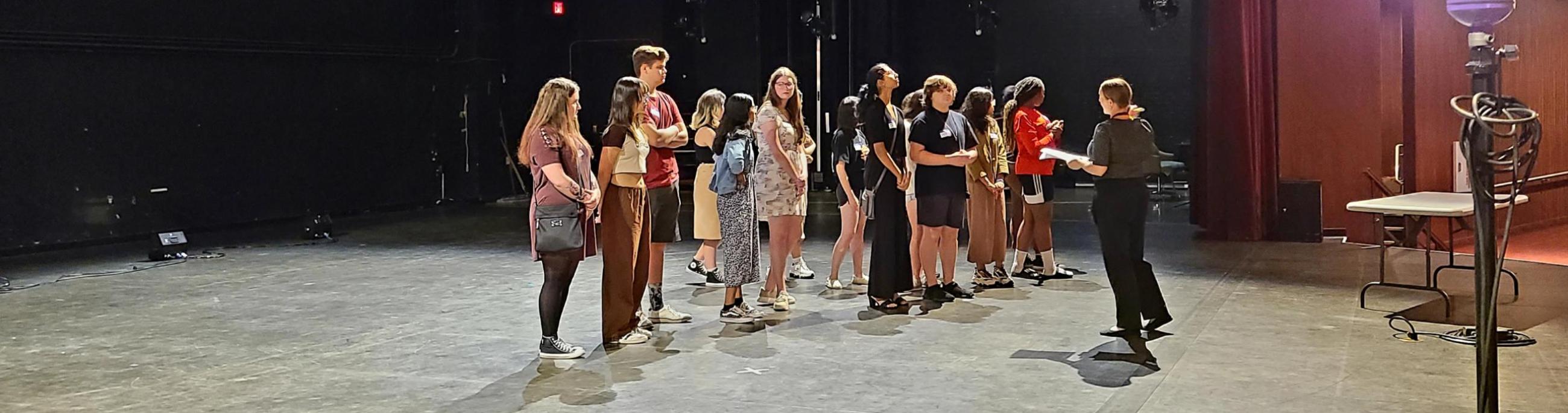 A group of students stands on a stage while a woman talks to them