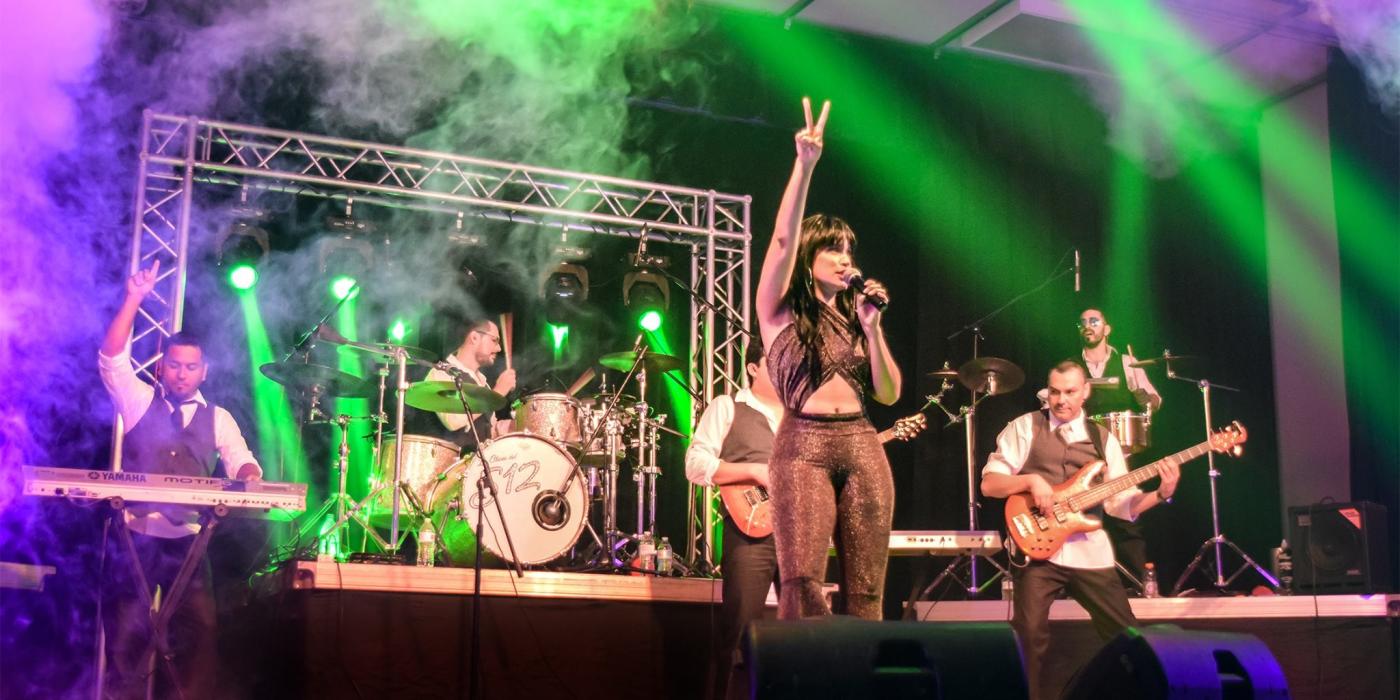 A full band plays on a stage with green and purple lights overhead. A woman stands at the front with one arm raised and a microphone in the other, singing.