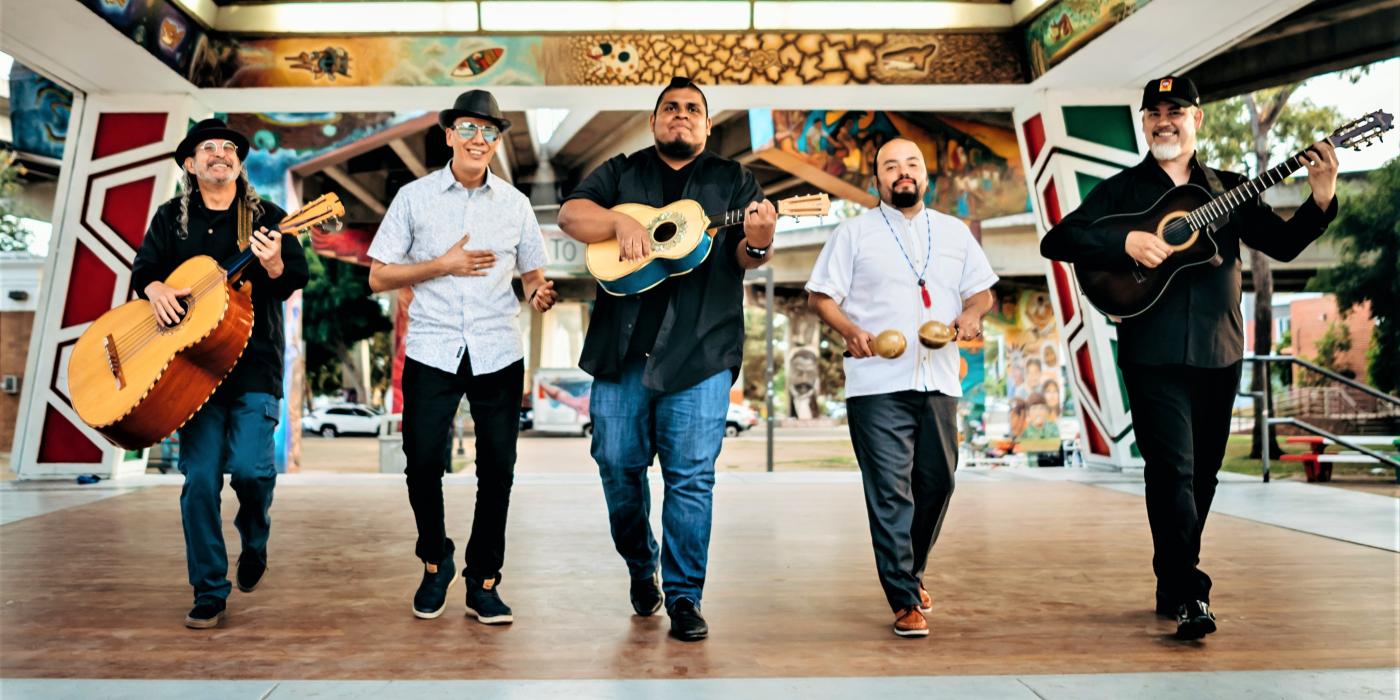 Five men walk through a courtyard toward the camera, four hold instruments