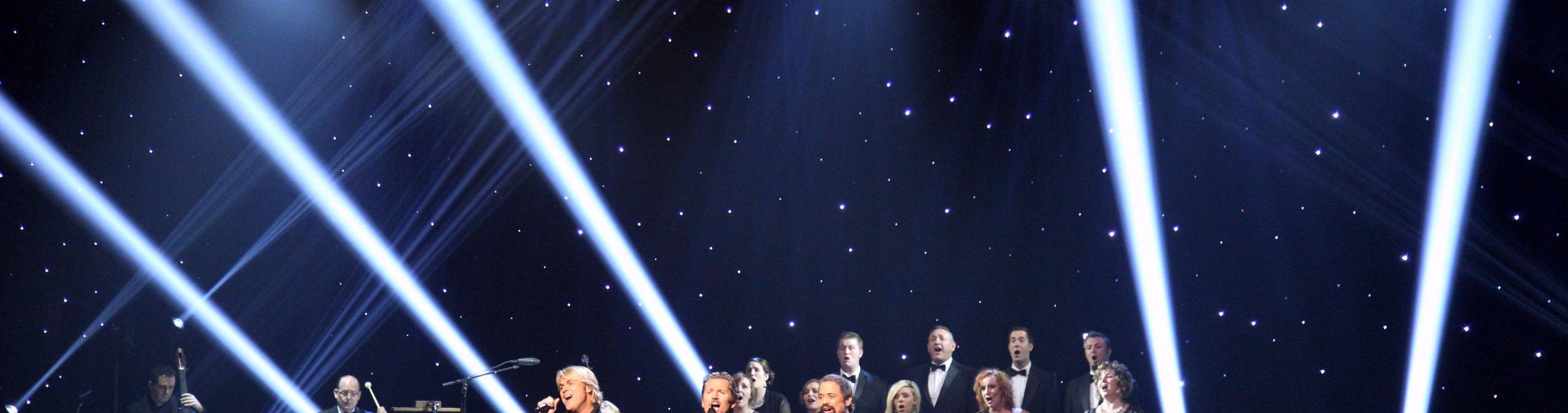 Three men with a chorus behind them standing on a dark stage singing