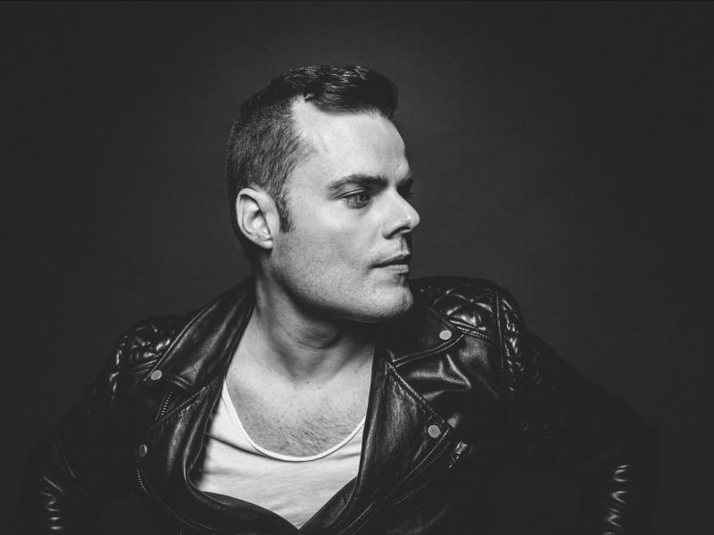 Black and white image of a caucasian male wearing a black leather jacket with white under-shirt.