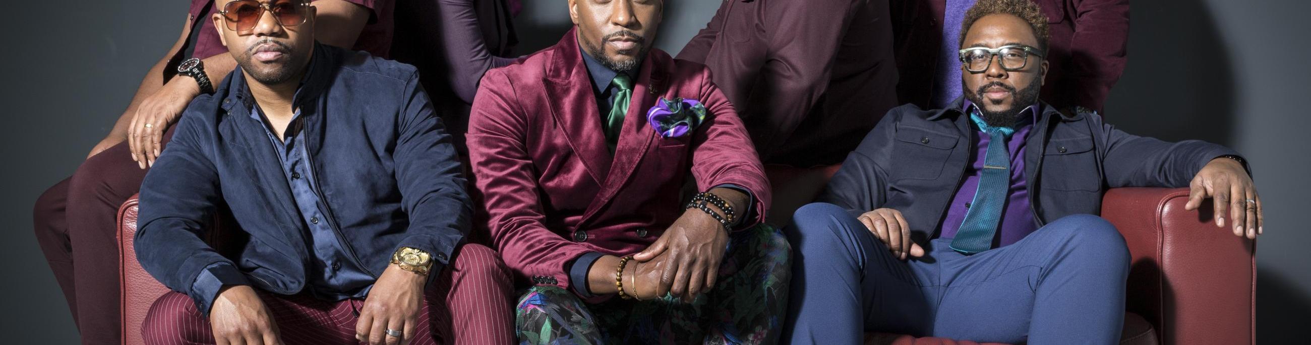 7 black men sitting around a maroon couch looking at the viewer