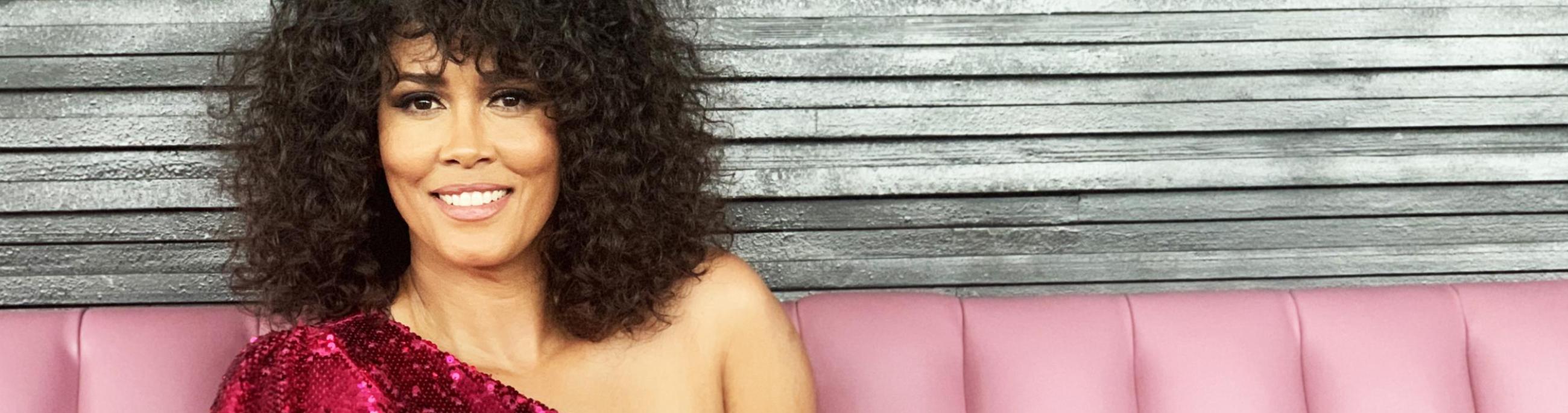 A woman with dark curly hair sites on a pink leather couch, wearing a magenta sequined top