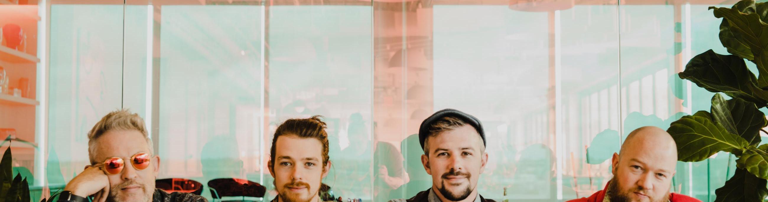 four white men sitting at a bar or table lined up and looking at the camera