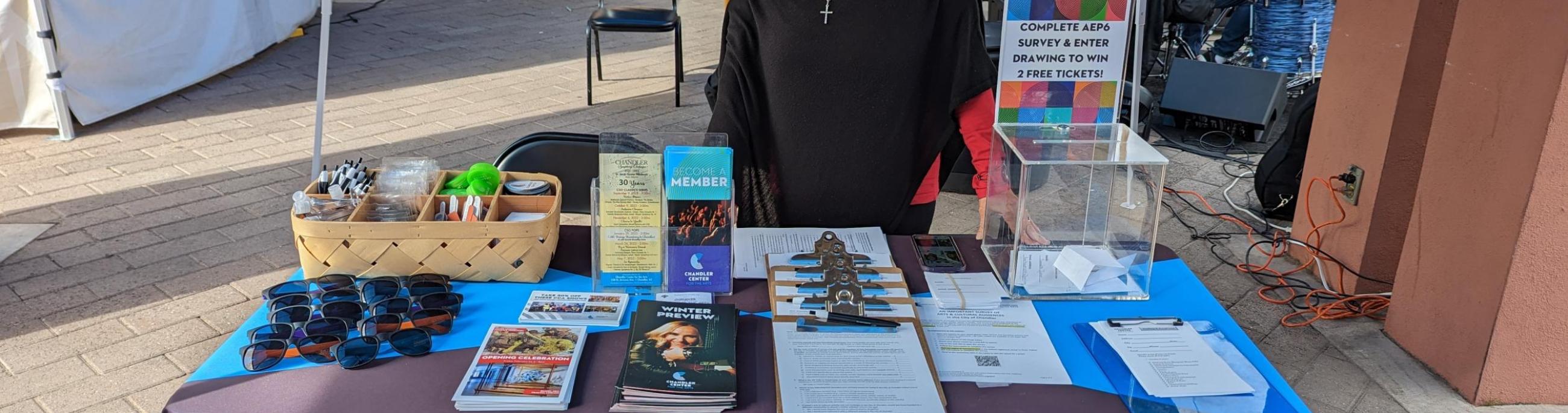 Woman in a black shirt and sunglasses standing behind a table with papers on it and a table cloth reading Chandler Center