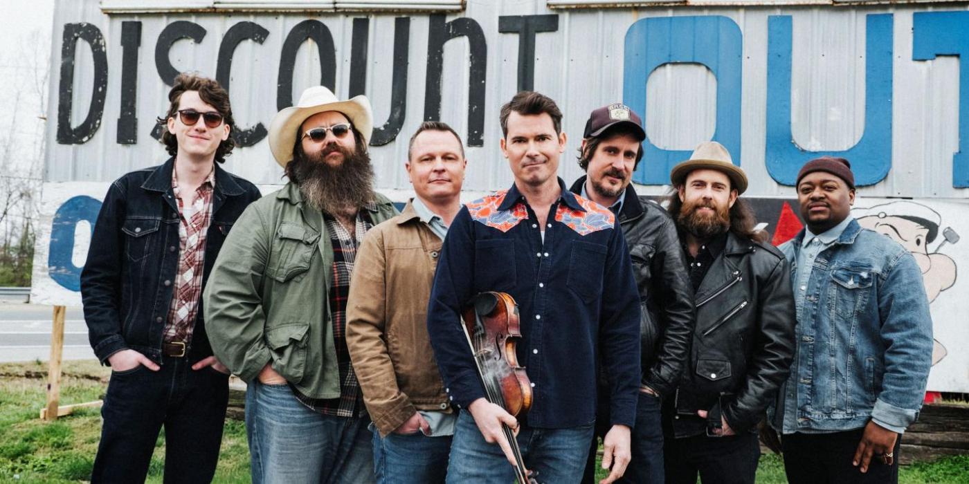 Seven men stand together in front of a sign advertising fireworks, wearing jeans and western shirts