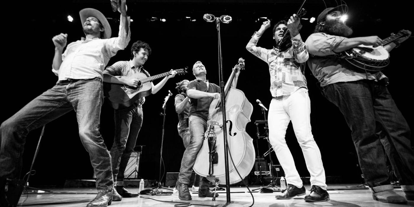 Four men dressed in jeans and traditional western wear hold string instruments while performing on stage