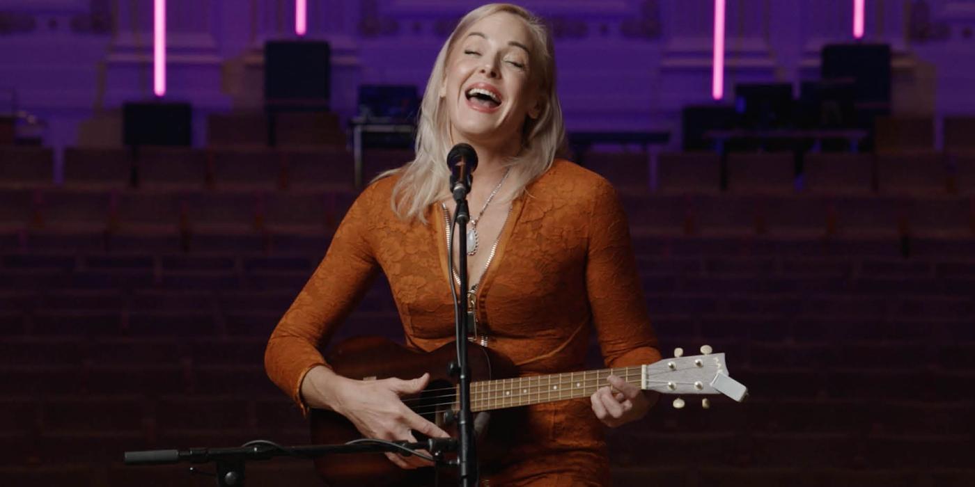 A woman sits on a tall stool in front of a microphone. Her mouth is open wide, eyes closed, singing out with a smile. She plays a ukulele.