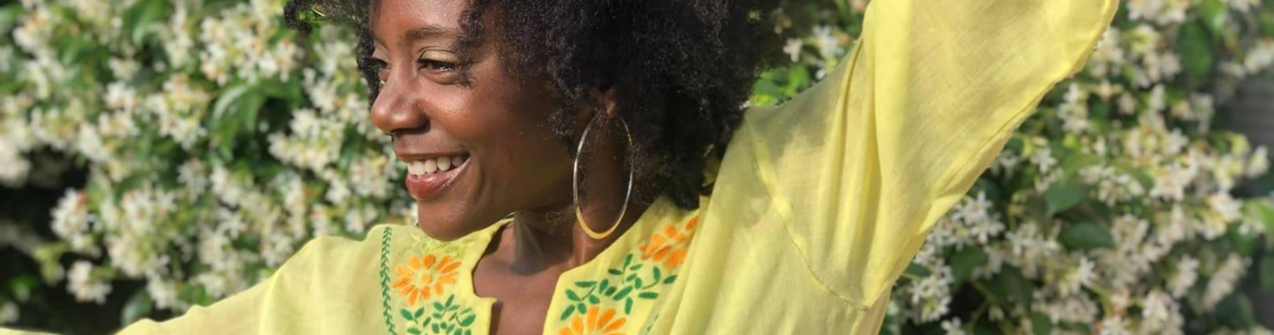 Niki J Crawford, wearing a bright yellow shirt, dances in front of a wall of white flowers.